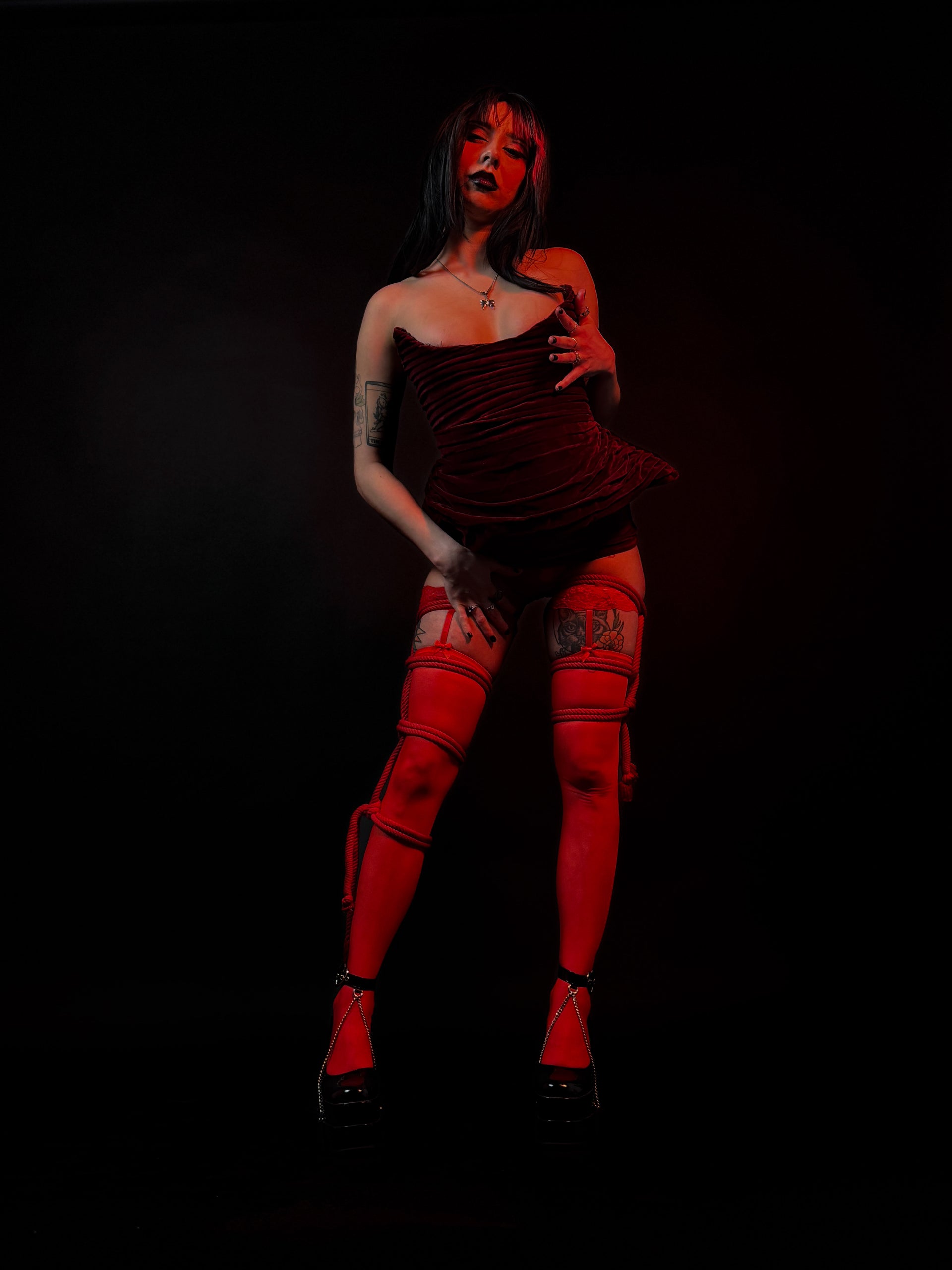 A model posing in a dark, strapless red dress, red thigh-high stockings with harness details, and red platform heels against a solid black background.